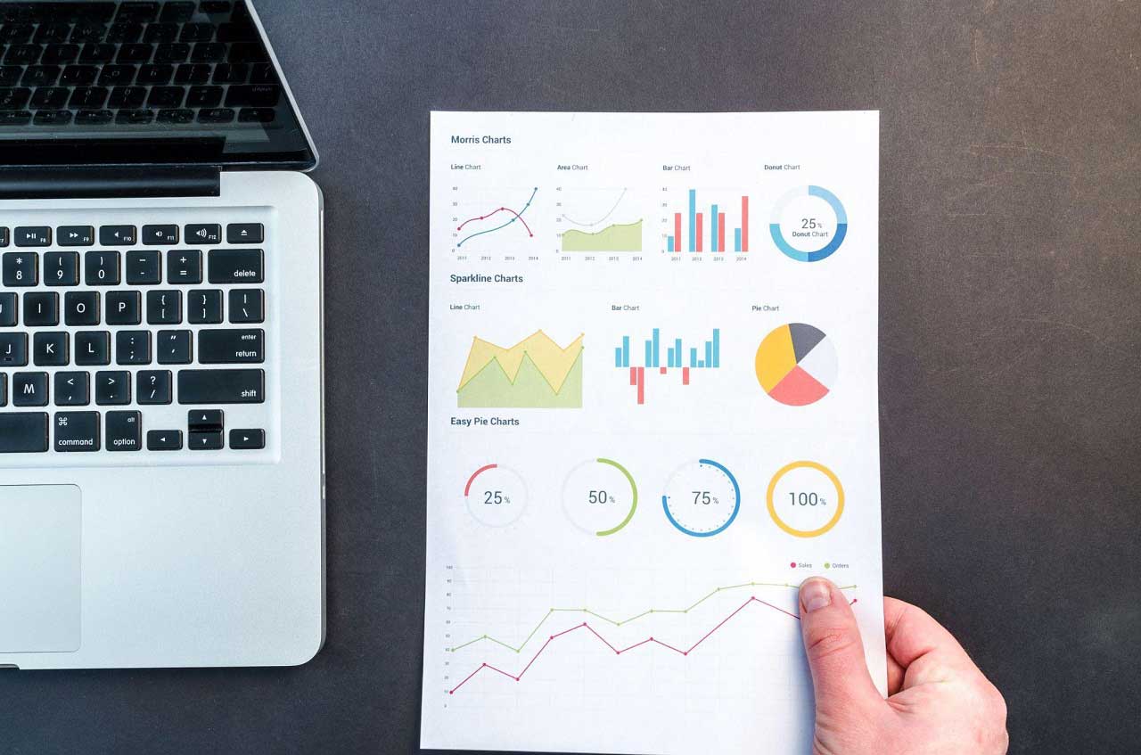 A desktop with a laptop on it and a person holding a paper with graphs.